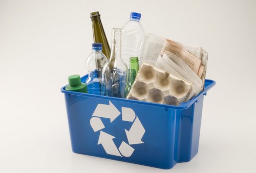 A blue plastic recycling container with a white recycling symbol on the front, filled with various waste items including glass bottles with blue and green caps, an empty clear plastic bottle, a cardboard egg carton, and crumpled newspapers. The container is placed on a plain white surface with a neutral background, highlighting the mixed recyclable rubbish suitable for removal or disposal services in Northolt. The glass bottles are transparent with different shapes and colors, and the newspapers are partially unfolded, adding to the visual detail of typical household waste. The scene emphasizes the collection of recyclable materials, consistent with rubbish removal activities provided by House Clearance Northolt in the local area.
