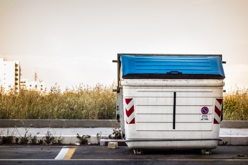 A large industrial wheelie bin with a blue lid positioned on a paved surface, situated at the edge of a parking area with a yellow line marking the space. The bin's white body features red and white reflective chevron strips on its sides and a small no parking sign attached to the front. Behind the bin, there is a strip of overgrown grass and weeds, with a backdrop of a blurred distant landscape that includes some trees and buildings, suggesting an urban or semi-urban setting in Northolt. The overall scene is illuminated by natural light, indicating daytime, and the setting appears to be an outdoor space likely associated with a property requiring waste collection or rubbish removal services, as handled by House Clearance Northolt.
