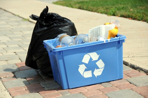 A blue plastic recycling bin with a white recycling symbol on its side is positioned on a paved sidewalk. The bin is filled with clear plastic bottles, some with labels, and a few have visible caps. Next to the bin is a full black trash bag, tied at the top, leaning against it. The background shows a grassy verge and part of a residential street, suggesting a typical street clearing or rubbish disposal scene. The setting appears to be outdoors in daylight, with natural light illuminating the scene, emphasizing the textures of the plastic bottles, the glossy surface of the bin, and the rough paving stones. This image is relevant to rubbish collection and waste management services, such as those offered by House Clearance Northolt, particularly in the context of household or local rubbish removal in Northolt or nearby areas.