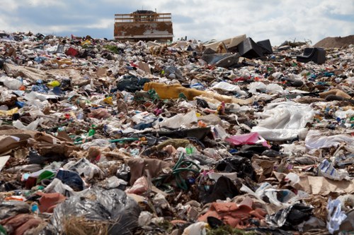 A vast open landfill site filled with a large accumulation of mixed household and commercial waste materials including plastic bags, cardboard, scrap paper, broken furniture, and miscellaneous rubbish, spread across a wide area with no clear organization. In the background, a partially constructed building or industrial structure is visible under a partly cloudy sky, indicating ongoing development nearby. The ground appears uneven and covered with debris, with no visible waste sorting or containment. Natural light highlights the textures and colors of the various types of rubbish, ranging from bright plastic items to duller cardboard and paper waste. This scene exemplifies the type of environmental challenge associated with improper waste disposal, relevant to professional rubbish removal services such as those offered by House Clearance Northolt in the Northolt area, serving local households and businesses in the region.
